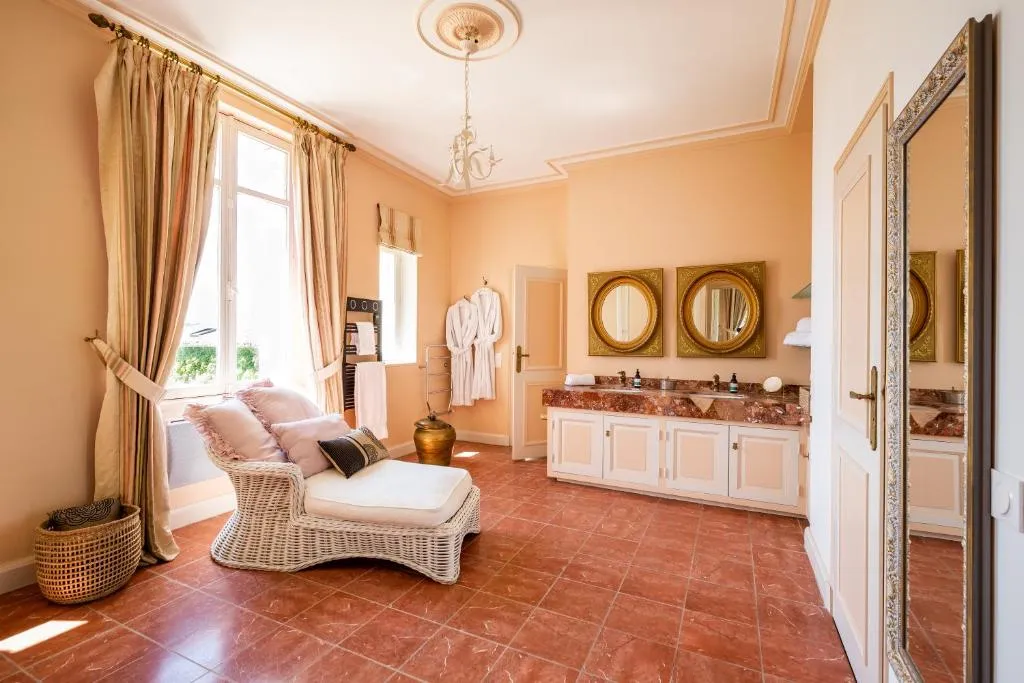 Salle de bain de la suite Colette avec meuble vasque en marbre rose, miroirs dorés et chaise longue en osier.